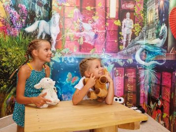 Zwei Kinder sitzen an einem Tisch vor einer farbenfrohen Wand in der Magischen Abenteuerwelt des Familienhotels Kaiserhof in Tirol, aufgenommen im Bereich Magische Bibliothek.