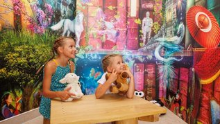Zwei Kinder sitzen an einem Tisch vor einer farbenfrohen Wand in der Magischen Abenteuerwelt des Familienhotels Kaiserhof in Tirol, aufgenommen im Bereich Magische Bibliothek.