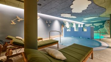 Innenbereich des Kinderpools im Zauchenseehof mit Liegen, spielerischer Burgwand, Felskulissen und Decke voller Wolken- und Vogelmotiven.
