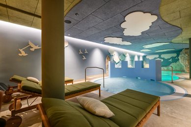 Innenbereich des Kinderpools im Zauchenseehof mit Liegen, spielerischer Burgwand, Felskulissen und Decke voller Wolken- und Vogelmotiven.