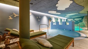 Innenbereich des Kinderpools im Zauchenseehof mit Liegen, spielerischer Burgwand, Felskulissen und Decke voller Wolken- und Vogelmotiven.
