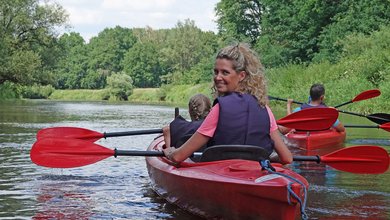 Frau und Kinder paddeln in roten Kajaks auf einem ruhigen Fluss im Emsland durch grüne Natur.