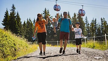 Eine Familie vom Wellness- & Familienhotel Egger geht den Erlebnisweg in Saalbach Hinterglemm spazieren, im Hintergund sieht man die Gondel 