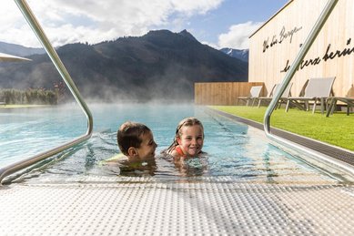 Zwei Kinder planschen im beheizten Außenpool des Familienhotels Almfamilyhotel Scherer mit Blick auf die umliegenden Berge.