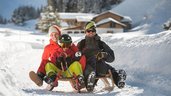Dreiköpfige Familie beim Schlittenfahren auf der familienfreundlichen Rodelbahn im Familienurlaub in Tirol, umgeben von einer verschneiten Winterlandschaft.