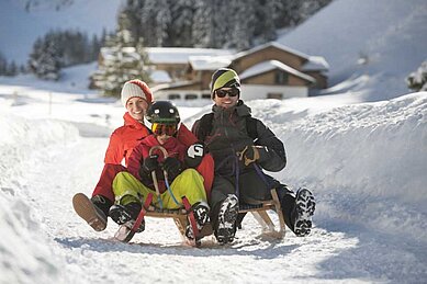 Dreiköpfige Familie beim Schlittenfahren auf der familienfreundlichen Rodelbahn im Familienurlaub in Tirol, umgeben von einer verschneiten Winterlandschaft.