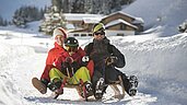 Dreiköpfige Familie beim Schlittenfahren auf der familienfreundlichen Rodelbahn im Familienurlaub in Tirol, umgeben von einer verschneiten Winterlandschaft.