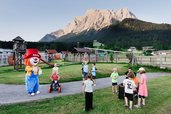 Kinder spielen draußen auf der Wiese mit dem Maskottchen Happy vor der Bergkulisse beim Familienhotel Tirolerhof.