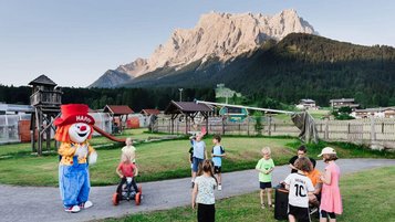 Kinder spielen draußen auf der Wiese mit dem Maskottchen Happy vor der Bergkulisse beim Familienhotel Tirolerhof.