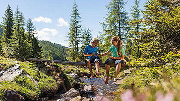 Zwei Kinder sitzen beim Wandern auf einem Baumstamm, der über einem Bach liegt.