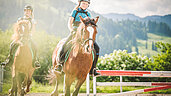 Zwei Kinder reiten auf zwei Pferden auf dem Reitplatz des Familienhotels Landgut Furtherwirt. Auf dem Reitplatz vom Landgut Furtherwirt gibt es jeden Tag die Möglichkeit für einen Ausritt oder eine Reitstunden. 