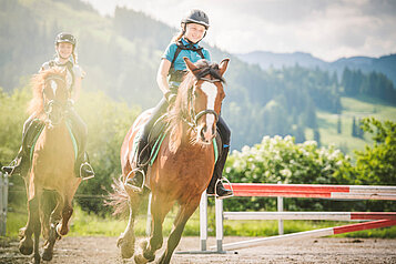 Zwei Kinder reiten auf zwei Pferden auf dem Reitplatz des Familienhotels Landgut Furtherwirt. Auf dem Reitplatz vom Landgut Furtherwirt gibt es jeden Tag die Möglichkeit für einen Ausritt oder eine Reitstunden. 
