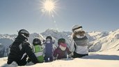 Familie sitzt in Skiausrüstung auf der Piste und ruht sich kurz aus, bevor es wieder ins Familienhotel Bella Vista in Südtirol geht.