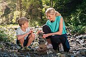 Mutter und Kind stapeln im Bach Steine und spielen in der umliegenden Natur des Familienhotels Das Hopfgarten in Tirol.