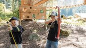 Zwei Kinder spielen Bogenschießen im Waldspielbereich des Familienhotels Huber in Südtirol