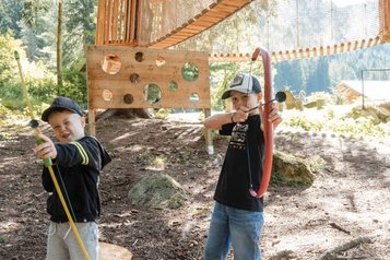 Zwei Kinder spielen Bogenschießen im Waldspielbereich des Familienhotels Huber in Südtirol