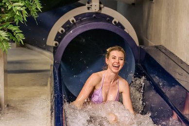 Eine lachende Frau in einem lilafarbenen Bikini rutscht aus einer dunklen Röhren-Wasserrutsche in ein spritzendes Wasserbecken und genießt sichtbar den Moment voller Spaß.