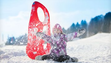 Fröhliches Kind sitzt lachend im Schnee und wirbelt Schneeflocken auf, neben ihm steht ein roter Rutschteller beim Familienurlaub im Winter.