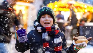 Ein fröhlicher Junge genießt die winterliche Stimmung auf dem Weihnachtsmarkt mit einer dampfenden Tasse und einer leckeren Bratwurst im Brötchen, während Schneeflocken in der Luft tanzen