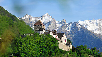 Liechtenstein im Sommer: Das Schloss Vaduz thront auf einem Berg, im Hintergrund verschneite Berge.