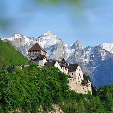 Liechtenstein im Sommer: Das Schloss Vaduz thront auf einem Berg, im Hintergrund verschneite Berge.