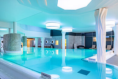 Entspannendes Hallenbad im Hotel Feldberger Hof mit blau leuchtendem Wasser, modernen Säulen und bequemen Liegestühlen in einer ruhigen Atmosphäre.