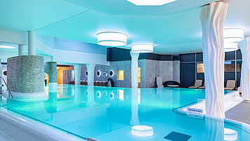 Entspannendes Hallenbad im Hotel Feldberger Hof mit blau leuchtendem Wasser, modernen Säulen und bequemen Liegestühlen in einer ruhigen Atmosphäre.