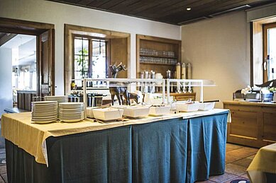 Einladendes Buffet-Setup in einem Hotelrestaurant mit einer Auswahl an Schalen und Tellern auf einer gedeckten Tischdecke, bereit für die Gäste, sich mit Köstlichkeiten zu bedienen.
