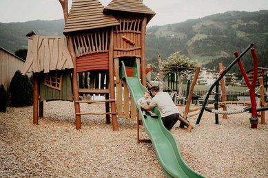 Ein Kind rutscht auf dem Spielplatz des Familienhotel Mia Alpina in Tirol neben Kletterturm, Schaukeln und Bergen.