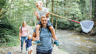 Familie mit zwei Kindern wandert im Wald am Fluss entlang. Die Kinder haben Kescher in der Hand.