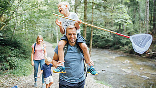 Familie mit zwei Kindern wandert im Wald am Fluss entlang. Die Kinder haben Kescher in der Hand.