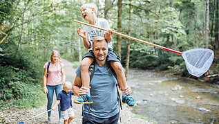 Familie mit zwei Kindern wandert im Wald am Fluss entlang. Die Kinder haben Kescher in der Hand.