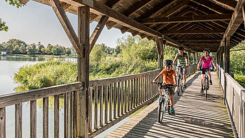 Zwei Eltern fahren mit ihrem Kind mit dem Fahrrad über eine hölzerne Brücke am See.