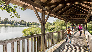Zwei Eltern fahren mit ihrem Kind mit dem Fahrrad über eine hölzerne Brücke am See.