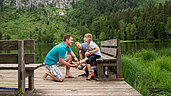 Mutter und Vater sitzen gemeinsam mit ihrer Tochter auf einer Bank am Frillensee in Bayern.