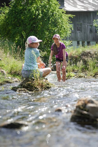 Zwei Kinder spielen lachend im Bach vor dem Familienhotel Sonne Bezau und spritzen mit Wasser.