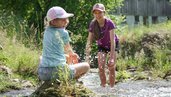 Zwei Kinder spielen lachend im Bach vor dem Familienhotel Sonne Bezau und spritzen mit Wasser.
