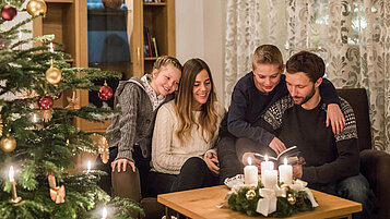 Winter im Salzburger Land: Familie sitzt zur Weihnachtszeit zusammen. Der geschmückte Christbaum im Vordergrund und auf dem Sofatisch steht ein Adventskranz.