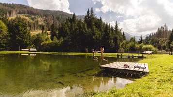 Zwei Kinder springen vom Steg in einen Natur-Badeteich am Almfamilyhotel Scherer in Tirol.
