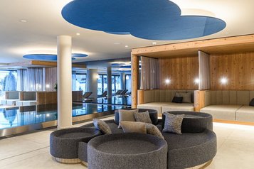 Moderner Ruhebereich im Zauchenseehof mit runden Loungeinseln, gemütlichen Polsterkojen und Blick auf den Innenpool, unter dekorativen Deckenelementen in Blautönen.