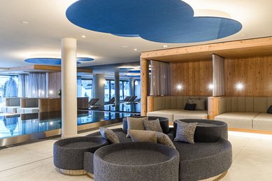 Moderner Ruhebereich im Zauchenseehof mit runden Loungeinseln, gemütlichen Polsterkojen und Blick auf den Innenpool, unter dekorativen Deckenelementen in Blautönen.