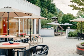 Sonnenterrasse mit runden Holztischen und Sonnenschirmen, im Hintergrund ein kleiner Spielplatz mit Holzrutsche und Bäumen im Familienhotel Amiamo.