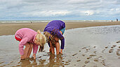 Zwei Mädchen buddeln ihre Füße im nassen Sand am Nordseestrand ein.