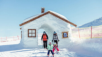 Dreiköpfige Familie fährt auf den Skiern durch einer aus Schnee gebauten Hütte.