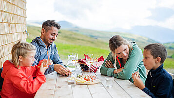 Familie mit zwei Kindern bei einer Pause von Wandern auf einer Hütte. Auf dem Tisch steht leckere Brotzeit.
