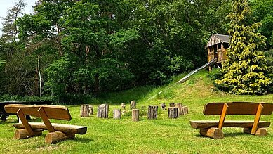 Sonniger Spielplatz mit Sandboden, Schaukeln und Volleyballnetz in einem familienfreundlichen Hotelgarten umgeben von grünen Bäumen, ideal für Freizeitaktivitäten im Freien.