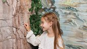 Ein Maedchen steht vor einer bemalten Wand mit Naturmotiv in der Magischen Abenteuerwelt des Familienhotels Kaiserhof in Tirol, aufgenommen im Bereich Feengrotte.