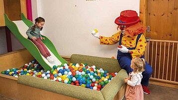 Kind rutscht auf einer Kinderrutsche ins Bällebad des Happy-Clubs im Familienhotel Alpenhotel Kind in Tirol. Auch Clown Happy, das Maskottchen des Familienhotels, spielt mit.