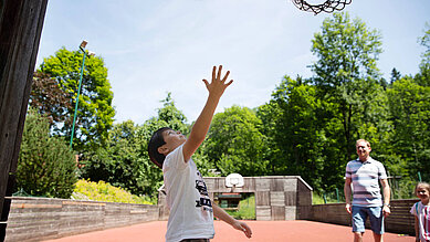 Jugendlicher Junge spielt mit seinem Vater Basketball auf dem Basketballplatz des Kinderhotels Bruckwirt.