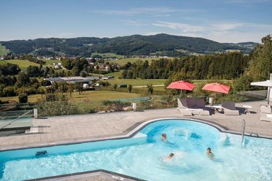 Außenpool im AIGO welcome family mit Badenden auf einer Terrasse, dahinter grüne Hügellandschaft und Felder unter blauem Himmel.